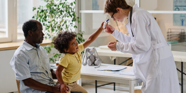 Female doctor trying to examine child with special medical equipment while he sitting with his dad at clinic